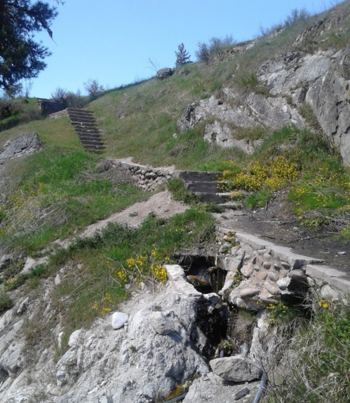 A rocky hillside with wooden steps leading up, surrounded by green grass and yellow wildflowers under a clear blue sky.